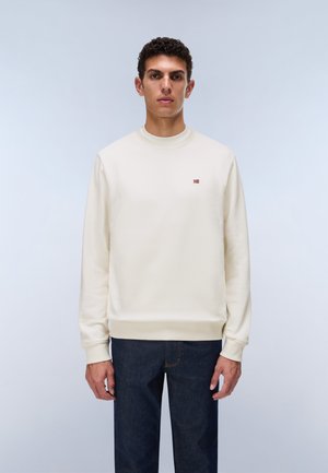 Jeune homme debout portant un sweat-shirt crème uni avec un petit emblème de drapeau rouge et blanc sur la poitrine et un jean bleu foncé.