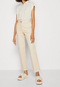 Beżowe bawełniane spodnie w połączeniu z lekką, pasiastą koszulą z krótkim rękawem i kremowymi sandałami typu slide z marszczonym detalem. Prosty fason, luźne dopasowanie.