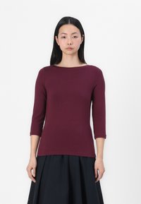 Burgundy tröja med figursydd passform, trekvartsärmar, rund halsringning och slät textur. Buren med en svart kjol, mot en enkel vit bakgrund.