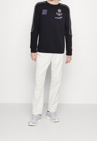 Sudadera negra con acentos blancos y varios parches coloridos, combinada con pantalones blancos y zapatillas gris claro. Look limpio y moderno.