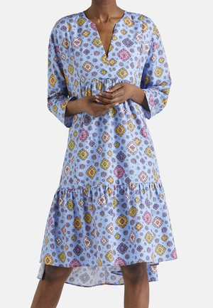 Kobieta ubrana w luźną, jasnoniebieską sukienkę z kolorowymi, diamentowymi wzorami kwiatowymi i długimi rękawami, stojąca ze splecionymi rękami.