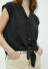 Schwarze kurzärmlige Bluse mit durchgehender Knopfleiste und gebundenem Saumdetail. Hergestellt aus einem glatten, leichten Stoff. Kombination mit beigen Hosen.