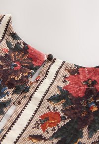 Gros plan d'une fermeture boucle de bouton en tissu avec un motif floral et géométrique en rouge, marron, noir et crème.