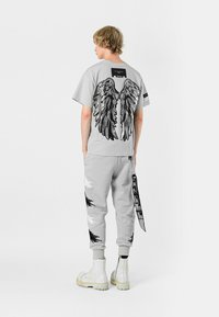 Camiseta gris con gráfico de alas en negro en la parte trasera, combinada con pantalones jogger grises que presentan acentos abstractos en negro y detalles en blanco, y botas blancas.