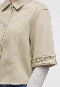 Chemise beige à boutons avec manches retroussées révélant une doublure intérieure à motifs, portée sur un haut gris clair à manches longues côtelé.