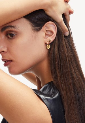 Goldohrringe mit einem Spiralmuster und einem runden Anhänger mit geometrischem Muster, getragen am Ohr eines Modells mit langen dunklen Haaren.