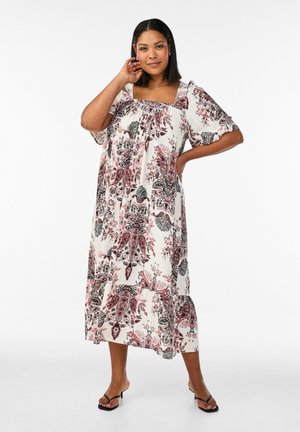 Woman wearing white midi dress with pink and black floral paisley pattern, square neckline, short sleeves, and black heeled sandals, posing.