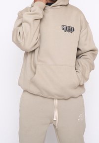 Béžová mikina s kapucí a přední kapsou, s nastavitelnou šňůrkou a logem. V kombinaci s béžovými tepláky, které mají boční kapsy a prošité detaily.