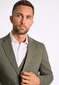 Olijfgroene blazer van een gestructureerde stof, met notch-lapel en een enkele borstzak. Draagt over een wit hemd met kraag.