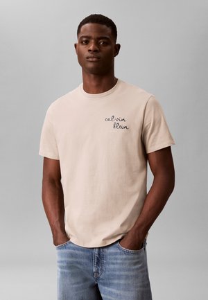 Bărbat tânăr purtând un tricou bej Calvin Klein și blugi albaștri, stând cu mâinile în buzunare, în fața unui fundal simplu gri.