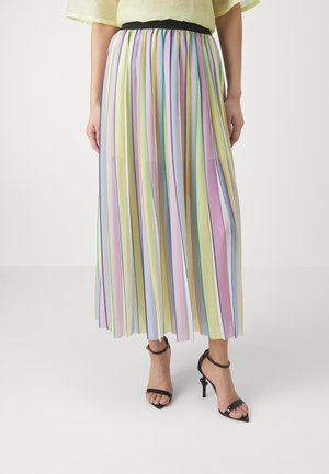 Femme portant une jupe midi plissée à rayures verticales pastel et des sandales noires à talons hauts, debout devant un fond uni.