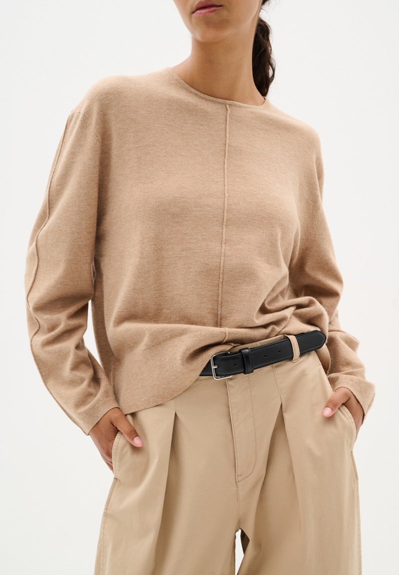 Maglione lavorato a maglia beige con collo a giro e maniche lunghe, caratterizzato da una cucitura centrale. Abbinalo a pantaloni beige a vita alta e una cintura nera.