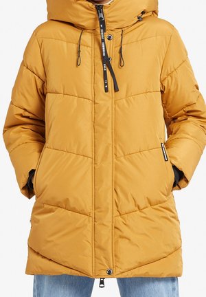 Personne portant une doudoune jaune moutarde avec une capuche, des cordons noirs, une fermeture éclair frontale et les mains dans les poches latérales.