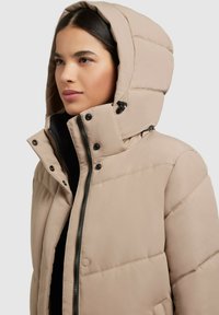 Jachetă puffer bej deschis cu glugă, având închidere cu fermoar, butoane cu capsenă și textură matlasată. Accente negre la fermoar și buzunare.