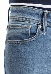 Blå denimjeans med hög midja, fem fickor och kontrasterande sömdetaljer. Har metallnitar och en mjuk, texturerad yta.