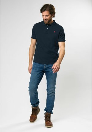 Camisa polo navy de manga corta, con cuello de canalé y un pequeño logo rojo. Combinada con unos jeans azules y botas de trabajo marrones.