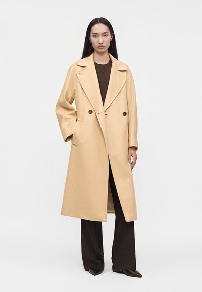 Femme portant un manteau long beige double boutonnage sur un haut foncé et un pantalon large, debout devant un fond clair uni.