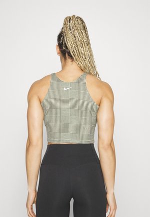 Osoba z długimi blond warkoczami, ubrana w wzorzysty krótki top i czarne legginsy z wysokim stanem, stoi tyłem na jednolitym tle.