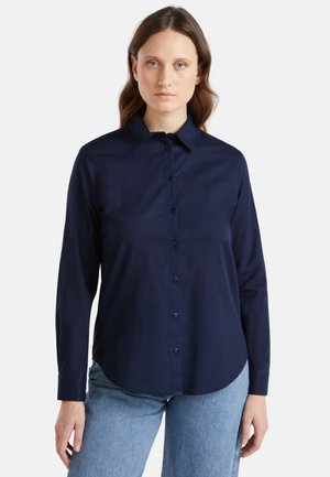 Donna dalla pelle chiara e capelli castani che indossa una camicia blu scuro con bottoni e jeans azzurri, in piedi davanti a uno sfondo bianco uniforme.