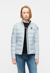 Νεαρός με κοντά κόκκινα μαλλιά, φοράει ανοιχτό μπλε μπουφάν καπιτονέ πάνω από μαύρο μπλουζάκι και μπλε τζιν, στέκεται μπροστά σε λευκό φόντο.