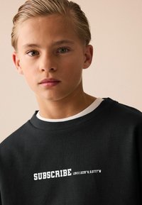 Sort sweatshirt med hvid tekst "SUBSCRIBE" og koordinaterne "LDN 51.5229° N 0.0777° W". Har en ribbet hals med et kontrasterende inderlag.