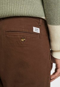Tasca dei pantaloni marroni con bottone e etichetta del marchio, abbinata a una manica e parte della mano visibili di un maglione lavorato a maglia verde e crema.