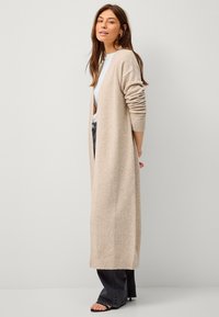 Beige langer Cardigan aus weichem Strickmaterial, mit offener Front, gerippten Bündchen und lockerer Passform, kombiniert mit dunkler Jeans und Sandalen.