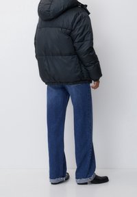 Sort puffjakke med hætte, brede blå jeans med flæser i kanten og sorte sko. Beklædningen har en quiltet tekstur og en afslappet pasform.