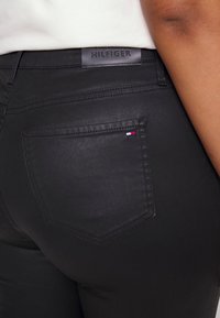 Detaljbild av svarta jeans med bakficka och liten Tommy Hilfiger-flagga på fickan samt läderetikett vid midjan.