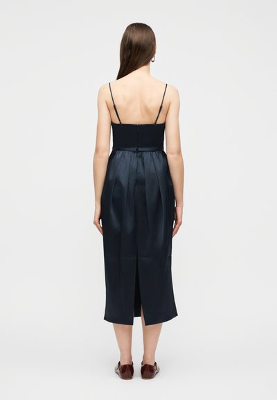 Γυναίκα με μακριά καστανά μαλλιά, φορώντας ένα μίντι φόρεμα χωρίς μανίκια σε σκούρο μπλε με ράχη και σκισίματα, και σκούρα κόκκινα φλατ παπούτσια, στέκεται σε λευκό φόντο.