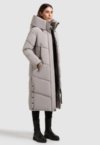 Kvinde iført en lang, lys grå puffjakke med høj krave, sorte leggings og sorte ankelstøvler, stående foran en ensfarvet baggrund.