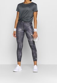 Grå atletisk t-shirt med knuten front, kombinerad med högmidjade leggings med geometriskt mönster i nyanser av grått, vita sneakers.