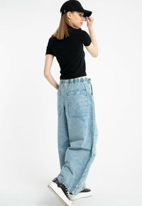 Kobieta w czapce, czarnym t-shircie, szerokich jasnoniebieskich dżinsach i czarnych trampkach, poprawiająca czapkę, stojąca na białym tle.