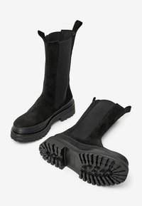 Černé semišové kotníkové boty s elastickými bočními panely, robustní gumovou podrážkou s texturovaným vzorem a poutky na horní části pro snadné obouvání.