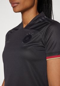 Czarna koszulka z fakturowanym wzorem, subtelnym kołnierzykiem i trzema paskami na ramionach. Akcenty w kolorze czerwonym i złotym na rękawach.