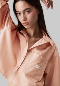 Camicia di cotone color pesca con vestibilità oversize, colletto ampio e tasca a toppa. Presenta cuciture a vista e orlo arrotondato.