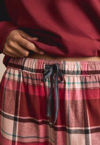 Punaiset ja vaaleanpunaiset ruudulliset pyjamahousut, joissa on joustava vyötärönauha ja musta kiristysnauha. Pehmeä kangas ruutu-urkuvalla ja kontrastiväreillä.