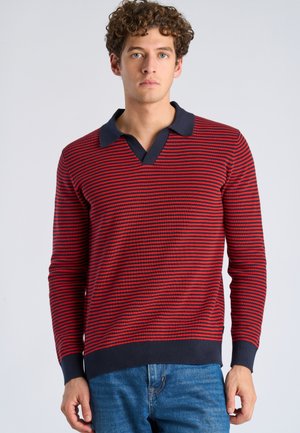 Ung mand med krøllet hår iført en rød og marineblå stribet langærmet polotrøje og blå jeans, stående foran en ensfarvet baggrund.