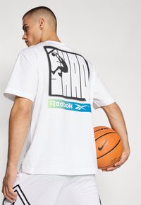 Bílý tričko s krátkým rukávem, na zadní straně s velkým černým grafickým potiskem a logem "Reebok" v zelených a modrých akcentacích. V ruce má basketbal.