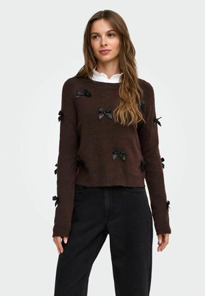 Femme portant un pull marron avec des nœuds en satin noirs, une chemise blanche à col en dessous, et un pantalon noir, debout avec les mains détendues le long du corps.