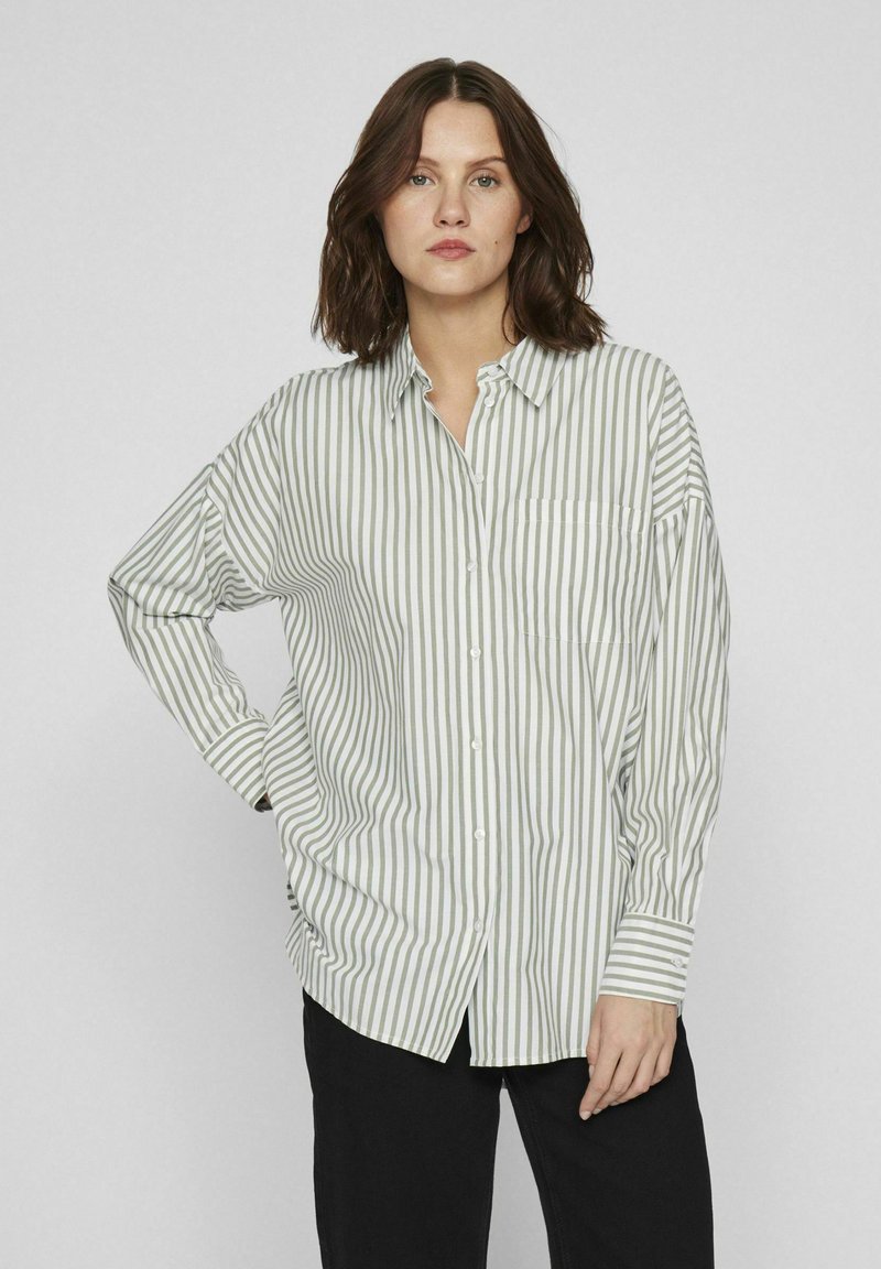 Camisa de botones con rayas verticales verdes y blancas, corte amplio, mangas largas, cuello y un bolsillo en el pecho.