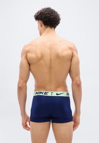 Άνδρας μοντέλο με σγουρά σκούρα μαλλιά, με μυώδη πλάτη, φοράει σκούρο μπλε εσώρουχα μπόξερ Nike με ανοιχτό πράσινο ζωνάκι, στέκεται μπροστά σε απλο υπόβαθρο.