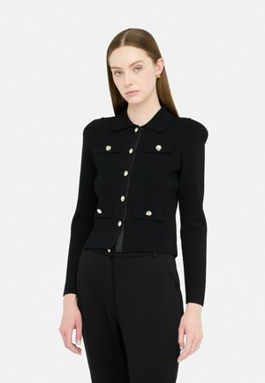 Femme portant une veste noire ajustée avec des boutons dorés et des poches, associée à un pantalon noir, se tenant devant un fond blanc.