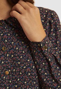 Blumenmusterhemd in Marineblau, mit kleinen roten, blauen und cremefarbenen Blumen, goldenen Knöpfen und mit Knöpfen versehenen Manschetten mit gefalteten Ärmeldetails.