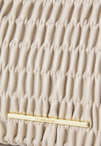 Beige lærveske med quiltet tekstur som har gullfarget aksent og preget logo. Mykt, fleksibelt materiale med fremtredende sømmer.