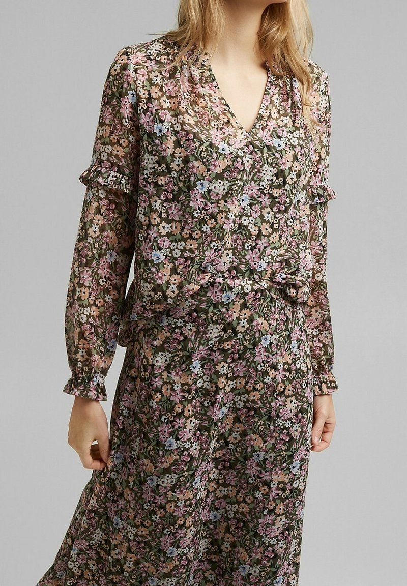 Femme portant une robe fleurie à manches longues avec un décolleté en V et des détails à volants sur les manches et la taille, debout devant un fond uni.