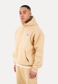 Mann trägt beige Hoodie und passende Jogginghose, steht vor einem schlichten hellen Hintergrund und blickt mit neutralem Gesichtsausdruck geradeaus.
