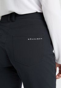Närbild av en person som bär svarta byxor med bakficka märkt "Röhnisch" och en vit långärmad topp.