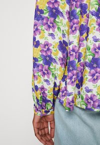 Main reposant à côté d'un jean bleu clair, portant une chemise à manches longues blanche avec un tissu imprimé floral en violet, jaune et vert.