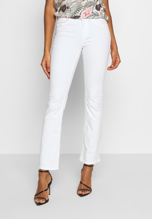 Kobieta ubrana w białe spodnie o prostych nogawkach, kwiecisty bezrękawnik i czarne sandały na wysokim obcasie z paskami, stojąca na tle jednolitej ściany.